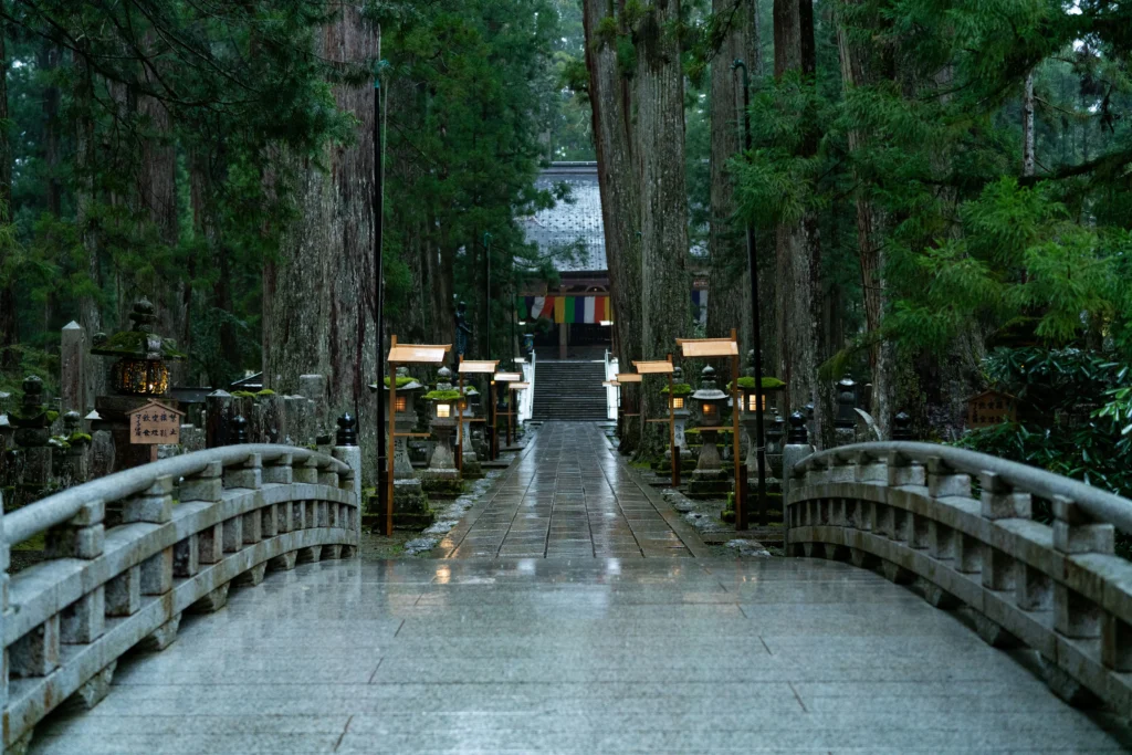 A wide, curved stone bridge wet from rain leads to a stone path lined with ancient cedar trees and glowing stone lanterns at Okunoin, Mount Koya. In the distance, a temple building with five-colored banners is nestled in the deep forest.