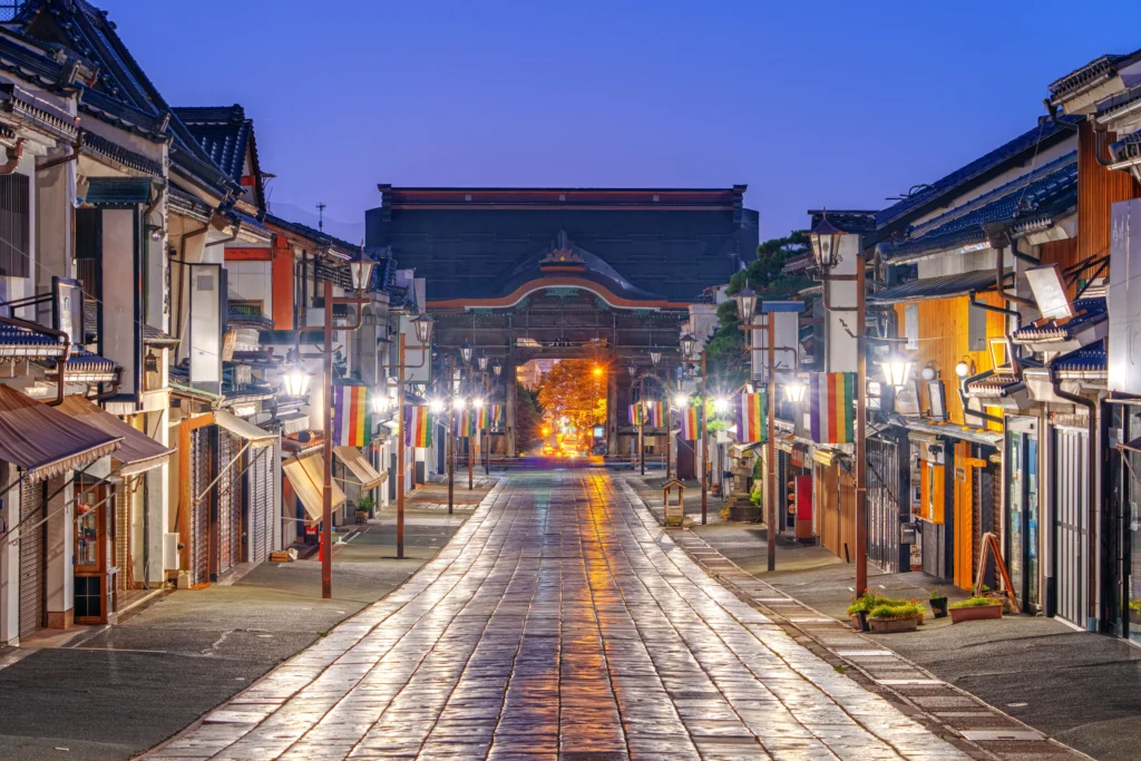 A tranquil view of the stone-paved approach to Zenko-ji Temple in Nagano at twilight. The wet pavement reflects the warm glow of street lanterns, and traditional buildings on both sides are decorated with colorful Buddhist banners (goshikimaku). The grand Niomon Gate stands at the end of the path under a deep blue sky.