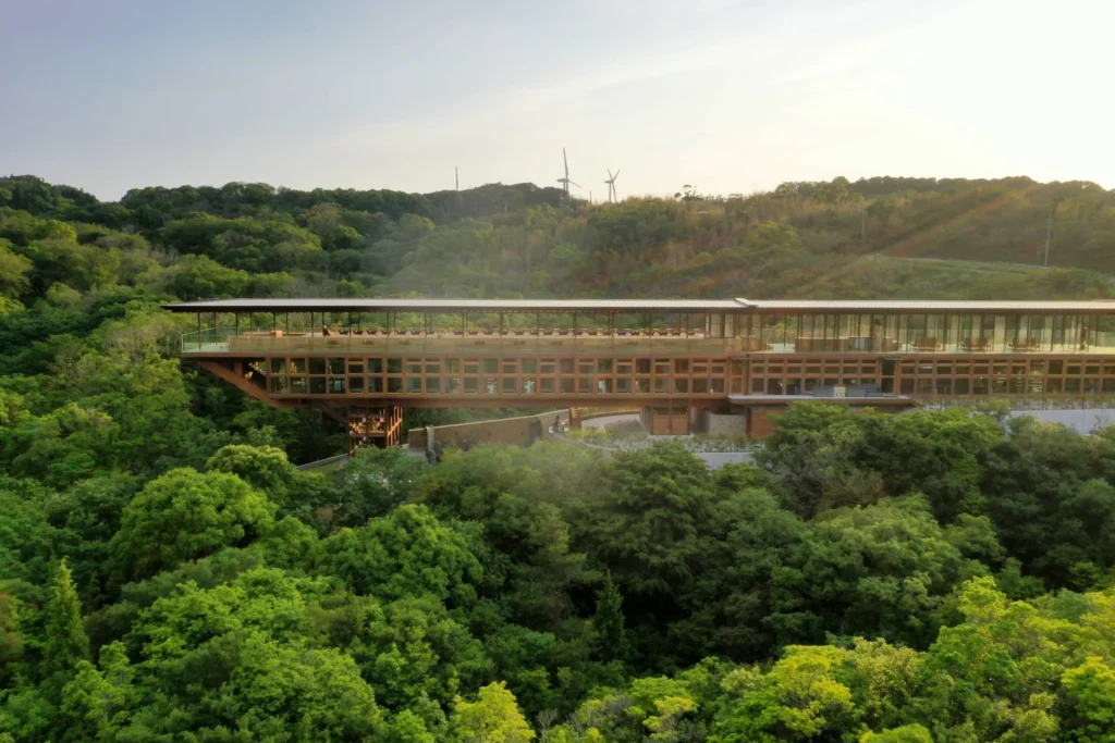 A wide aerial view of Zenbo Seinei, a long, slender wooden architecture perched on a verdant hillside on Awaji Island. The 100-meter-long bridge-like structure is nestled among a dense forest under a soft, hazy sky. Its repetitive wooden lattice and open deck seamlessly blend with the surrounding greenery, creating a sense of being suspended in nature.