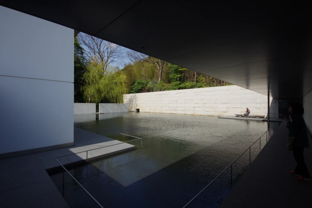 A view from the shaded corridor of the D.T. Suzuki Museum looking out towards the stone wall and the Water Mirror Garden. The dark foreground of the corridor frames the sunlit exterior, where a visitor sits on a wooden bench against a large, textured stone wall. Lush greenery and trees rise behind the wall under a clear sky.