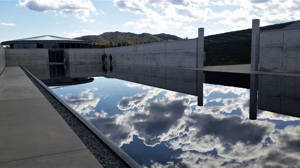 A serene wide-angle view from the exterior of the Head Buddha complex, looking across a large, still reflection pool. A minimalist concrete walkway flanks the left. In the distance, a circular, low building sits nestled in forested hills. A dramatic and perfectly clear reflection of the voluminous white clouds and blue sky dominates the pool's surface. Two visitors stand in the distance, providing a sense of scale against the concrete walls.