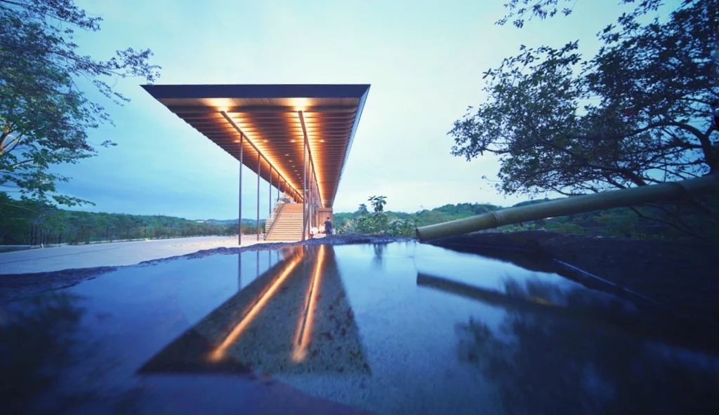 A low-angle evening shot focusing on the entrance of Zenbo Seinei. In the foreground, a traditional stone water basin (tsukubai) with a bamboo spout reflects the warm, glowing lights from the building's underside. The long, cantilevered wooden roof stretches toward the twilight sky, with a staircase leading up into the illuminated interior, evoking a serene and spiritual atmosphere.