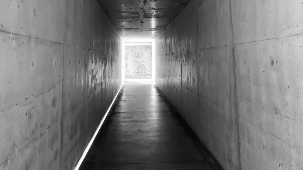 A symmetrical, minimalist view looking down a long, narrow concrete corridor inside the Chichu Art Museum. Smooth reinforced concrete walls line both sides, marked with a grid of formwork holes. At the far end, a rectangular opening frames a sunlit space where a large, abstract wall sculpture is partially visible.