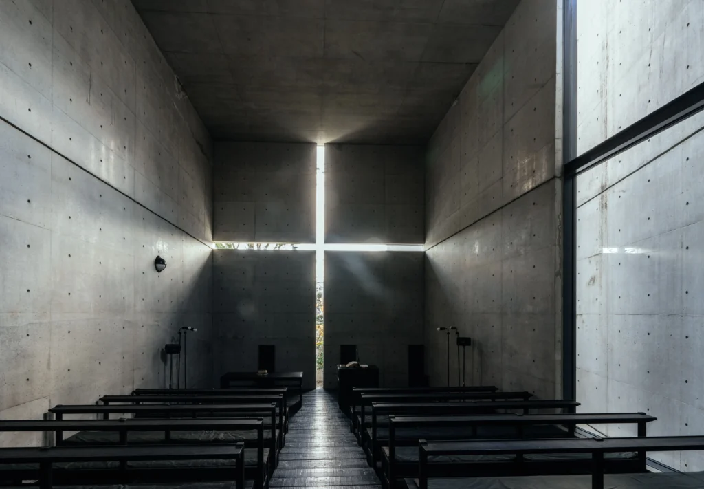 An interior view of the Church of the Light, a minimalist concrete chapel. Natural light streams through a cruciform (cross-shaped) opening cut into the front wall, illuminating the dark, atmospheric space. Rows of simple black wooden pews face the light, and the walls are made of smooth, unadorned reinforced concrete with a grid of circular tie-rod holes.