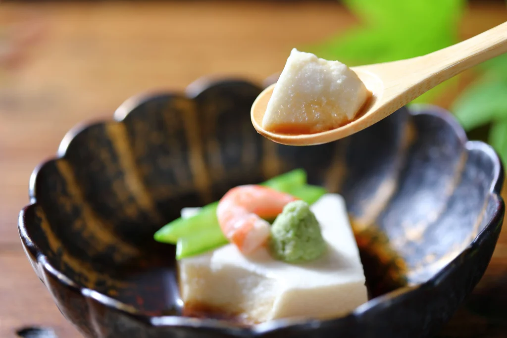 A close-up of a bite of silky white sesame tofu (Goma Dofu) on a wooden spoon, held over a dark ceramic bowl garnished with a shrimp, wasabi, and snap peas.