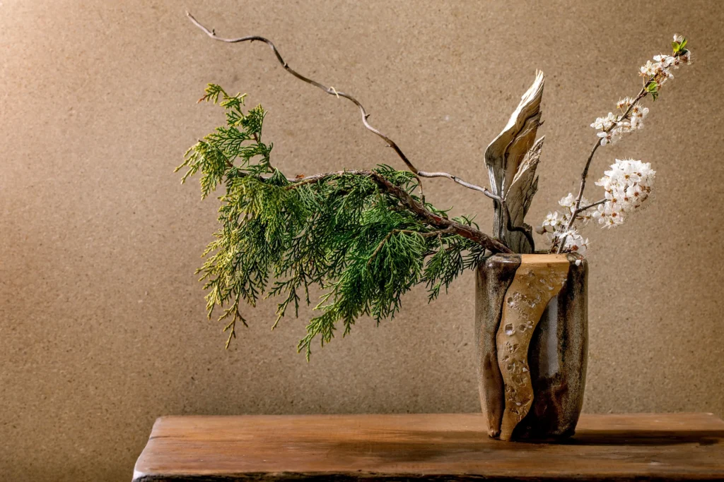 An Ikebana flower arrangement in a tall, textured ceramic vase, featuring green cedar branches, delicate white blossoms, and a stark, curved dry branch against a neutral wall.