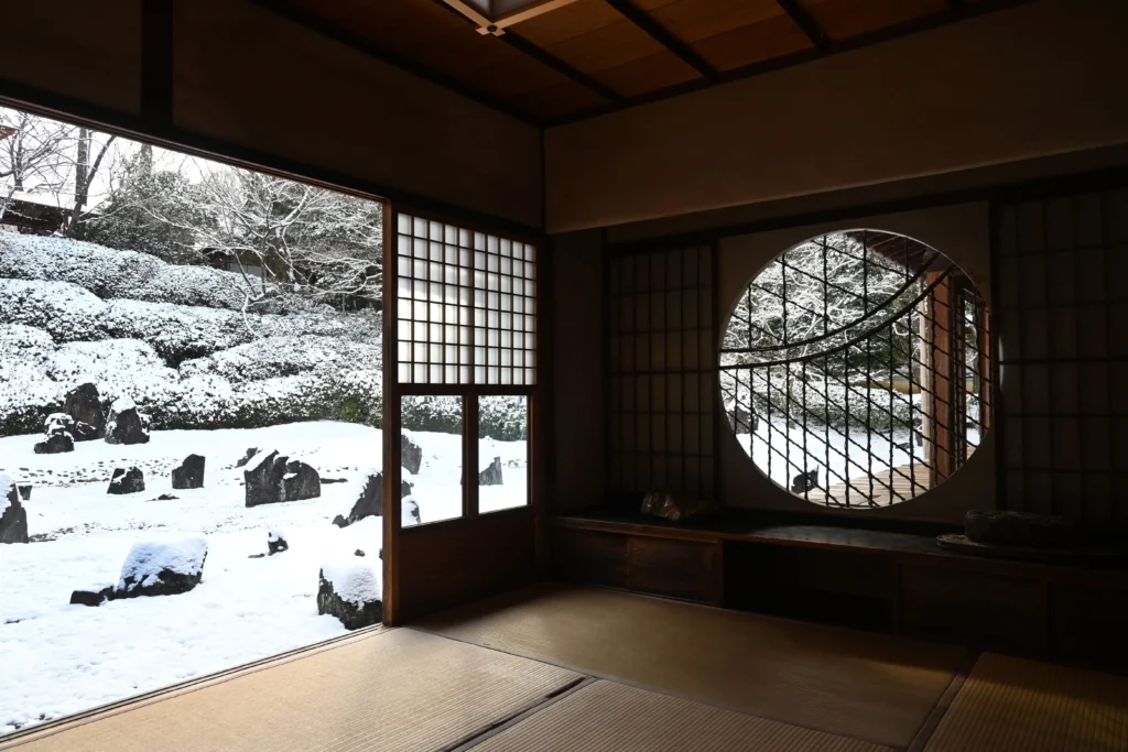 Interior of a traditional Japanese room with tatami mats, featuring a large open sliding door and a circular "Marumado" window that frame a Zen rock garden covered in a thick blanket of white snow under a grey winter sky.
