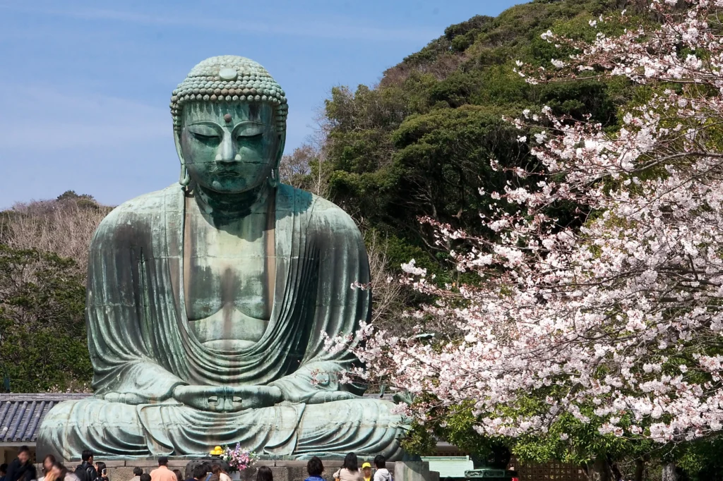 The Kamakura Great Buddha (Daibutsu) at Kōtoku-in—the iconic bronze statue seated in the open air, framed by trees, with visitors providing scale. Clear sky, warm light.