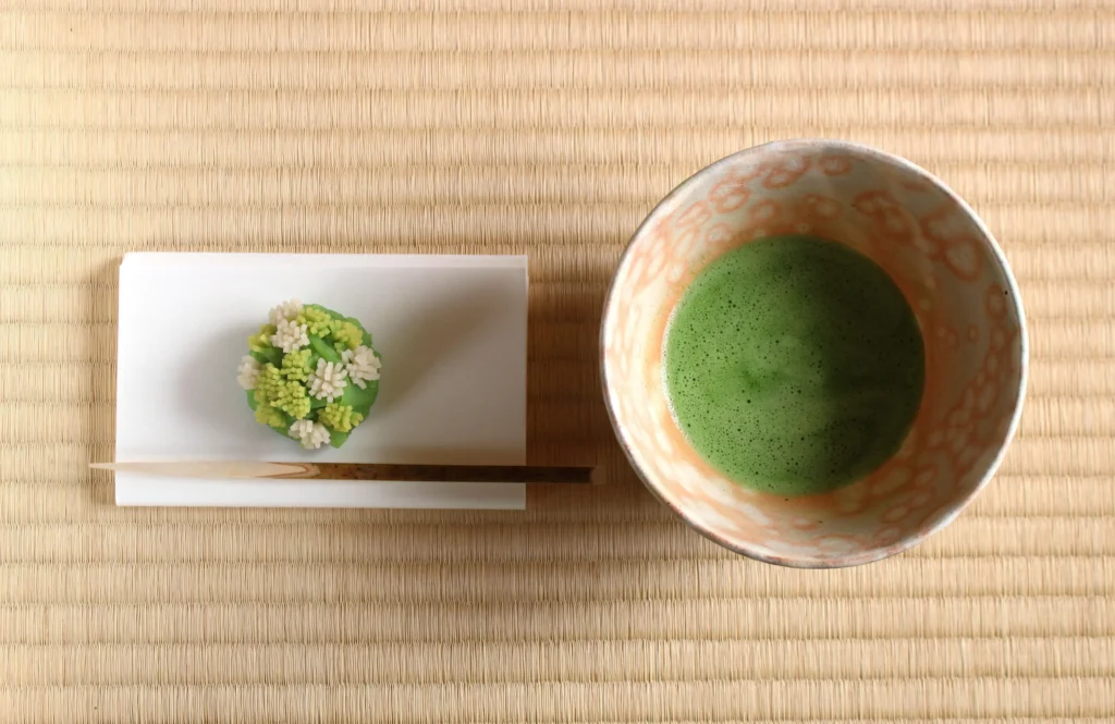 A top-down view (flat lay) on a tatami mat, showing a bowl of frothy green matcha tea next to a delicate green and white wagashi sweet served on white kaishi paper.