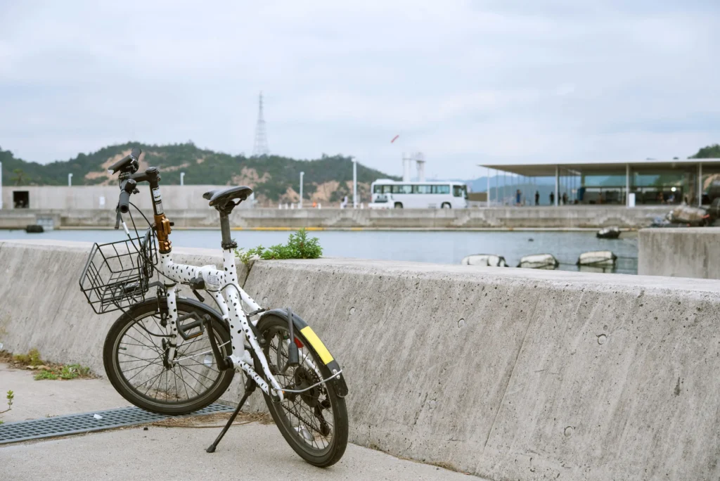 A white rental bicycle with black polka dots parked against a concrete sea wall at Miyanoura Port in Naoshima. In the background, the sea, a white bus, and the minimalist architecture of the ferry terminal are visible under a cloudy sky.
