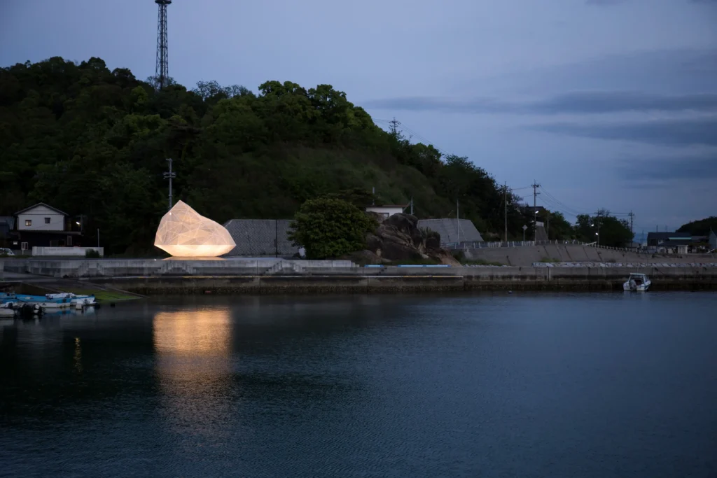 Night view of the Naoshima Pavilion, a glowing white polyhedral mesh structure by Sou Fujimoto, located at Miyanoura Port. The illuminated artwork is reflected on the calm dark water of the harbor against a backdrop of a forested hill at dusk.