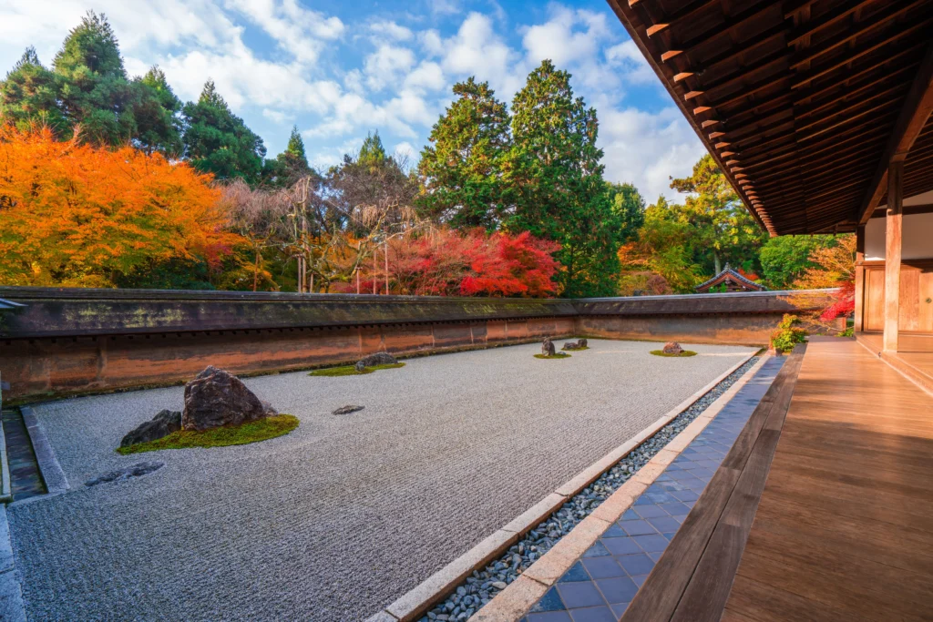 A wide-angle view of the Ryoanji Zen rock garden in Kyoto from a wooden veranda. The garden features meticulously raked white gravel and islands of rock and moss, surrounded by a low earthen wall. Vibrant red, orange, and green autumn foliage rises behind the wall under a bright blue sky with white clouds.