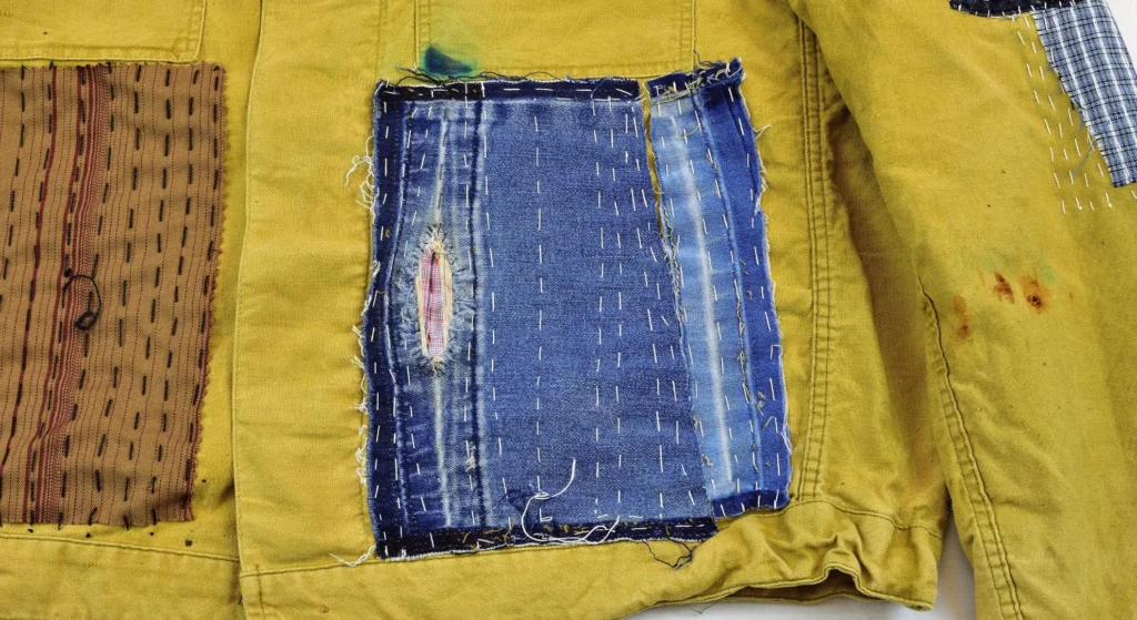 A close-up of a mustard yellow canvas jacket featuring decorative visible mending. Several patches of denim and striped fabric are hand-stitched onto the garment using white and dark thread in a traditional Japanese sashiko style, creating a rustic, textured aesthetic.