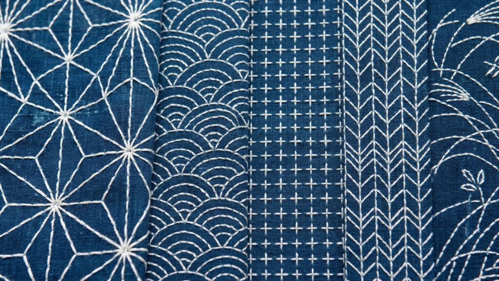 A horizontal composition of five indigo fabric panels, each featuring a different traditional white Sashiko pattern. From left to right: the star-shaped "Asanoha" (hemp leaf), the rhythmic "Seigaiha" (ocean waves), the geometric "Jūjitsunagi" (linked crosses), the directional "Yabane" (arrow feathers), and the flowing "Nowaki" (autumn grasses). Each panel showcases the contrast between the deep blue textile and the precise hand-stitched motifs.