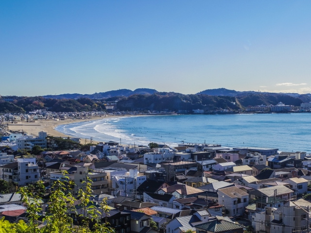 An elevated, wide-angle view from Hasedera Temple overlooking the coastal city of Kamakura. The scene features a cluster of residential rooftops in the foreground leading to the crescent-shaped sandy beach of Yuigahama Bay. Gentle waves roll onto the shore under a clear blue sky, with lush green hills surrounding the coastline in the distance.