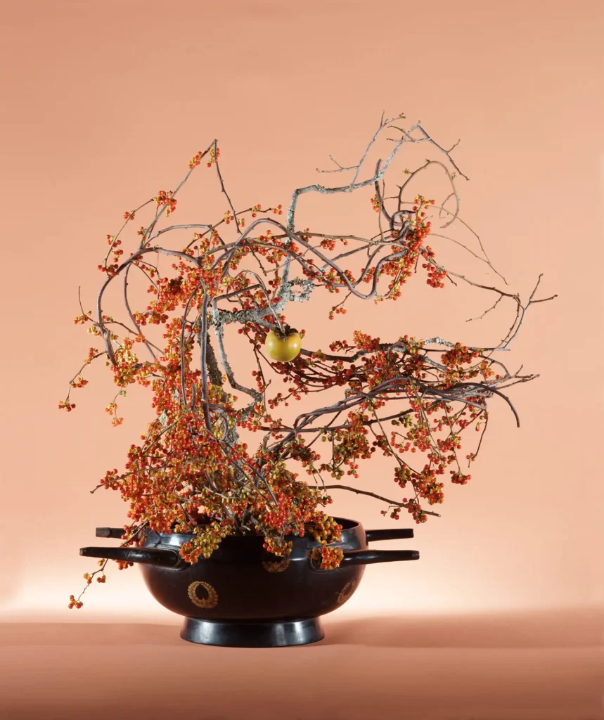A dynamic and sculptural Sogetsu-ryu Ikebana arrangement in a large black ceremonial vessel with handles. The arrangement features a dense, swirling mass of branches laden with small red-orange berries, with a single green persimmon hanging as a focal point in the center, set against a warm peach background.