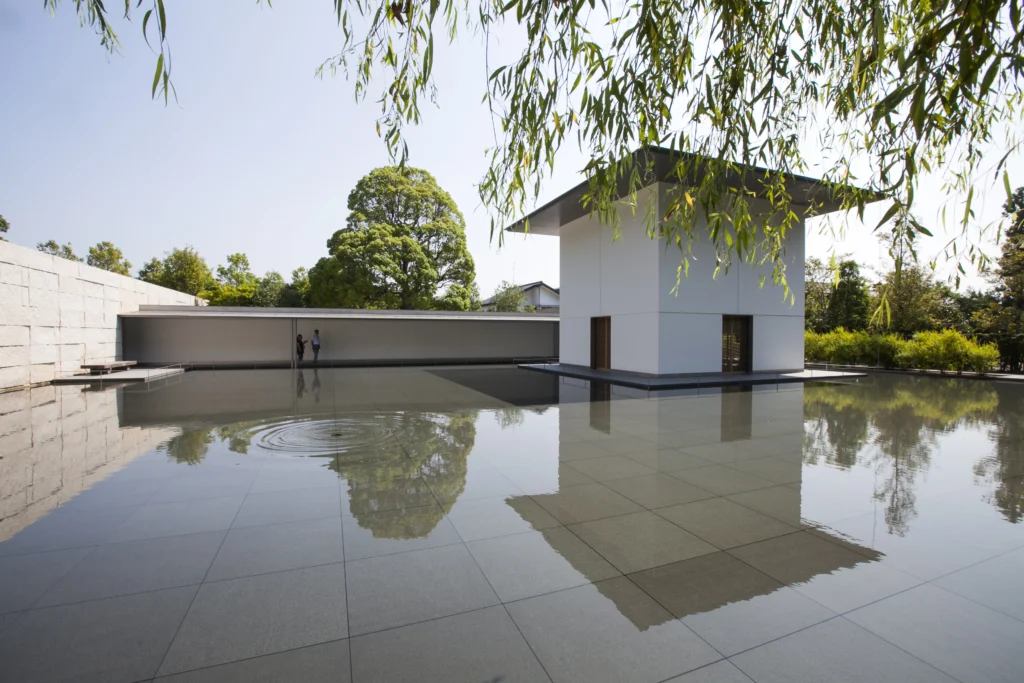 A bright, wide-angle view of the "Water Mirror Garden" at the D.T. Suzuki Museum. A minimalist white cubic structure, the Contemplative Wing, stands in a shallow reflecting pool. The still water creates a perfect mirror image of the building and the sky, with gentle ripples forming in the foreground. Willow branches hang softly from the top of the frame.