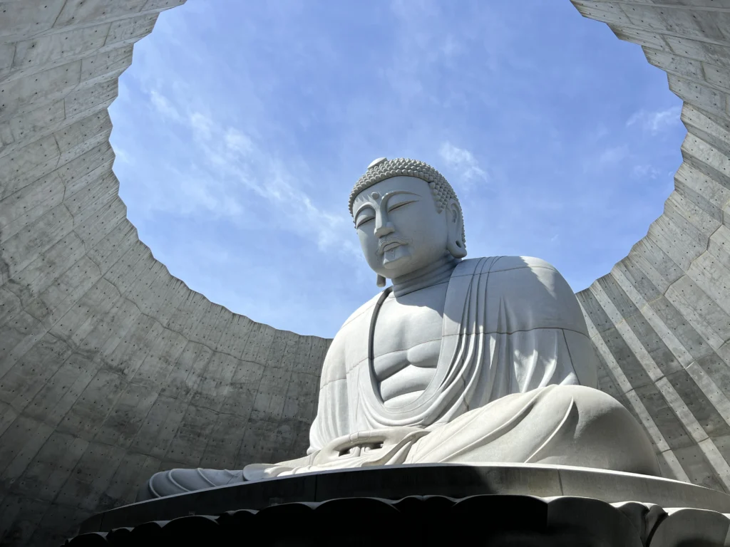 An awe-inspiring low-angle shot looking up at the Atama Daibutsu (Head Buddha). The massive stone statue, seen from below, sits serenely within a modern, circular concrete structure. The concrete 'halo' perfectly frames a view of the bright blue sky and wispy white clouds above the statue's serene, partially sun-drenched face.