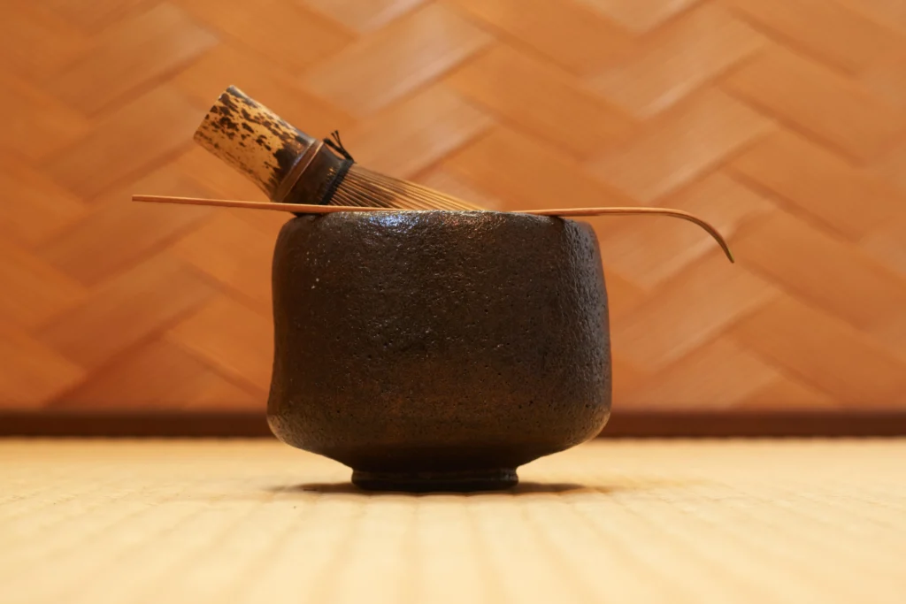 A dark brown Raku-ware tea bowl (chawan) sitting on a tatami mat with a bamboo whisk (chasen) and tea scoop (chashaku) resting on top.