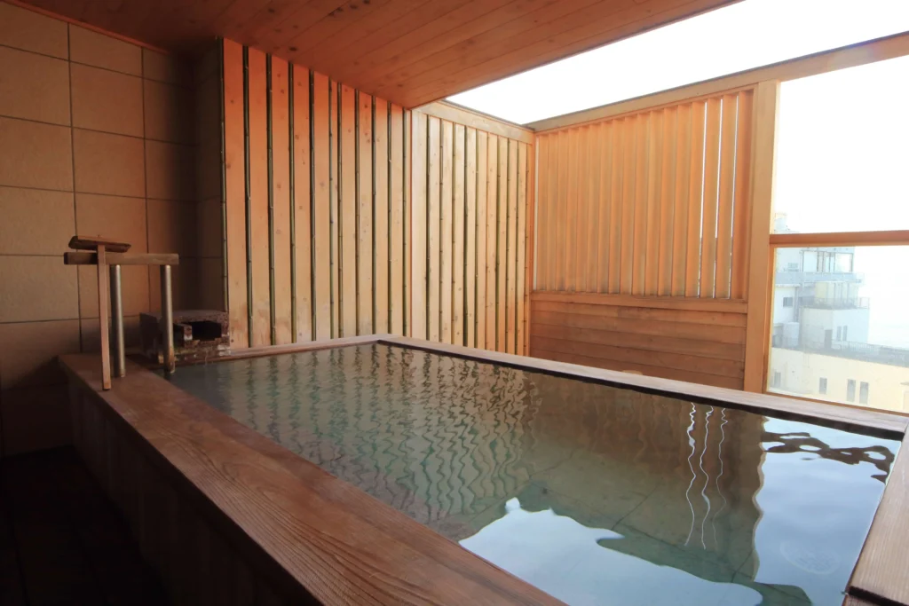 A private indoor hot spring bath (kashikiriburo) featuring a rectangular wooden tub filled with clear water, surrounded by bamboo-textured walls and large windows letting in natural light.