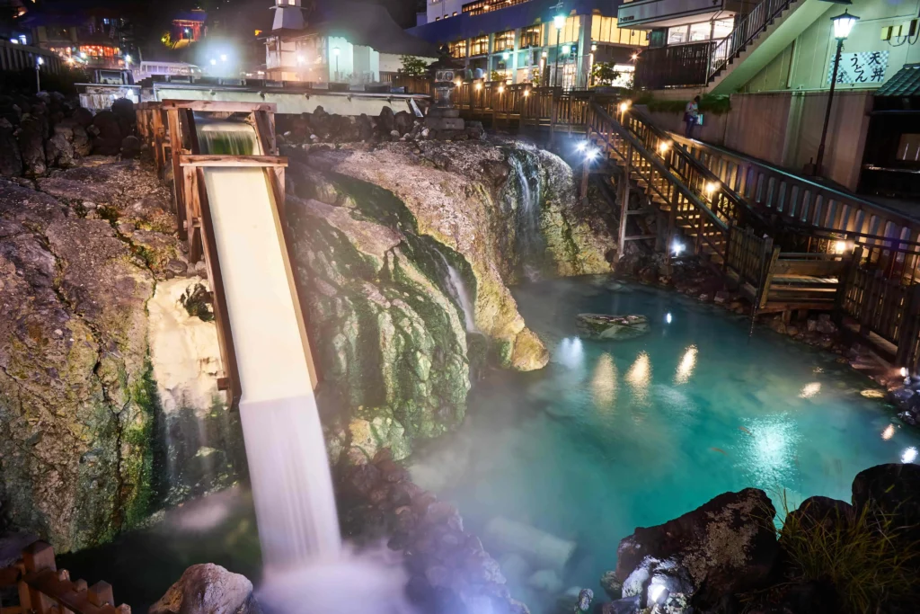 Night view of Kusatsu Onsen's Yubatake (hot water field), with milky white water flowing down a large wooden chute into a steamy, vibrant blue-green pool. The area is illuminated by warm lights, showing stone-paved walkways and multi-story Japanese buildings.