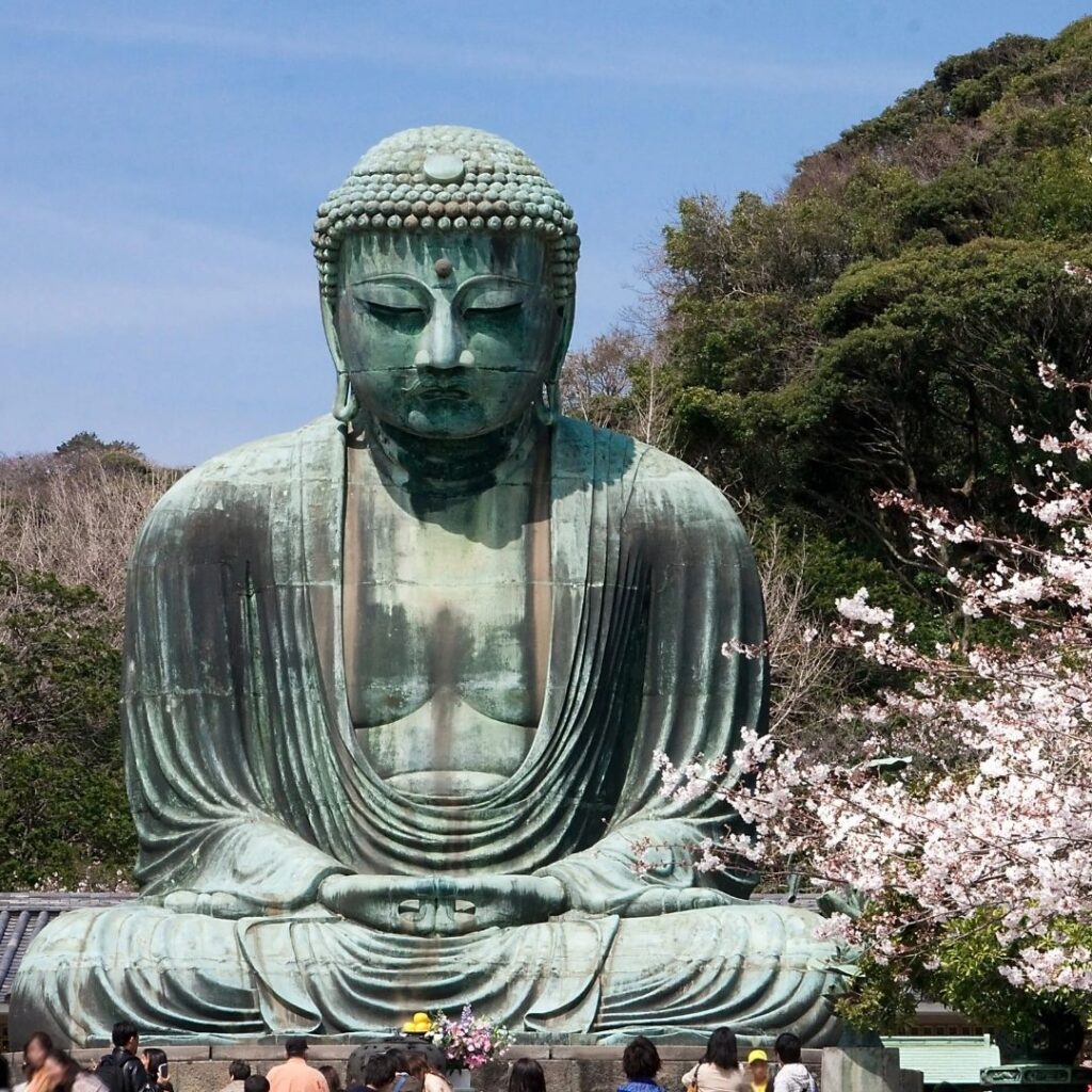 The Kamakura Great Buddha (Daibutsu) at Kōtoku-in—the iconic bronze statue seated in the open air, framed by trees, with visitors providing scale. Clear sky, warm light.