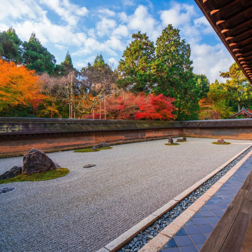 A wide-angle view of the Ryoanji Zen rock garden in Kyoto from a wooden veranda. The garden features meticulously raked white gravel and islands of rock and moss, surrounded by a low earthen wall. Vibrant red, orange, and green autumn foliage rises behind the wall under a bright blue sky with white clouds.