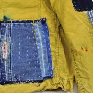 Top-down detailed photograph of a distressed mustard-colored canvas jacket. It features multiple artisanal patches: a large central panel of layered, frayed blue denim and a side panel of vertical brown striped fabric. Both are meticulously secured with prominent white sashiko running stitches and dark thread, showing the visible signs of a well-loved and repaired garment.