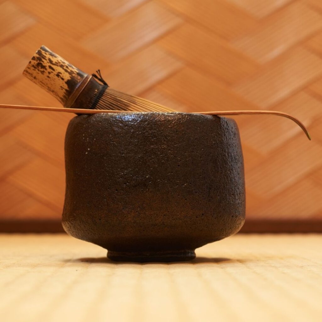 Close-up of a handmade Black Raku tea bowl on a straw tatami floor, featuring a rustic texture and traditional bamboo tea utensils.